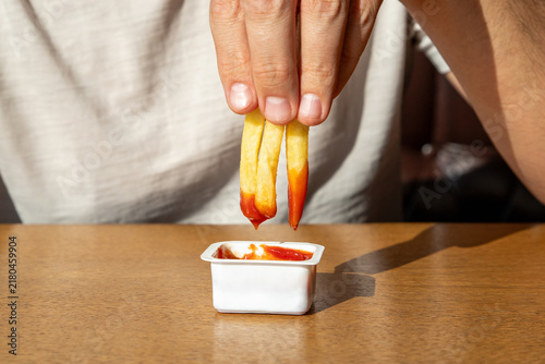 Man dipping french fries into container with ketchup. Close up of male hand holding fried potato sticks over sauce. Fast food snack and unhealthy eating concept for cafe menu and food advertising
