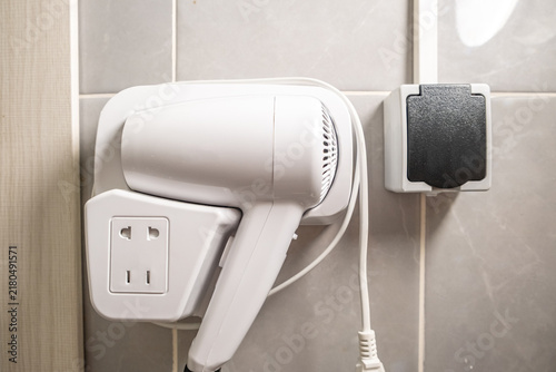 Hair dryer mounted on wall in hotel bathroom with electrical outlet and power switch, featuring modern design and sleek white finish against tiled background