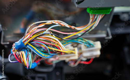 ​A close-up bundle of colorful electrical wires with plastic connectors hangs from a disassembled car dashboard. The auto electronics repair process shows complex cable wiring during onboard system ma