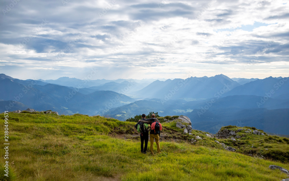 Obraz premium Two climbing friends with ropes attached to their backpacks looking across the Bavarian Alps, scenic mountain panorama, alpine adventure, outdoor friendship, active travel and shared exploration