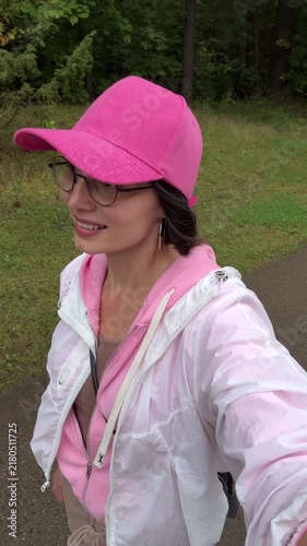 Inside profile view of a cheerful young woman in a pink baseball cap and eyeglasses walking outdoors in a park and smiling during a fresh morning walk.