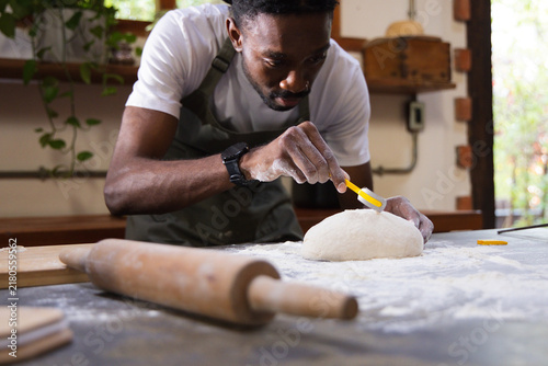 Canvas Print African man wearing gray apron leaning over floured counter scoring dough with y