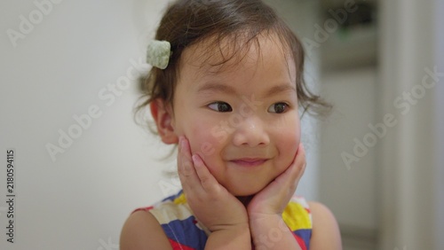 Фотография A cute Asian toddler girl holds her face with both hands and smiles gently, looking away with a sweet expression of childhood wonder and contentment in her home