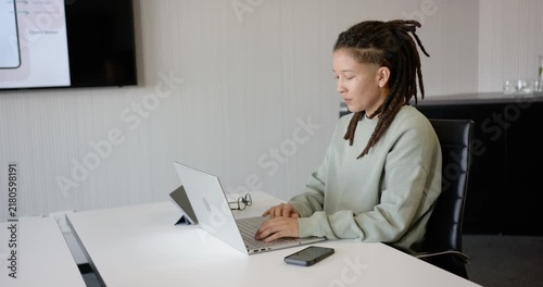 Nonbinary African American typing laptop at long white meeting table, glancing at slide, copy space