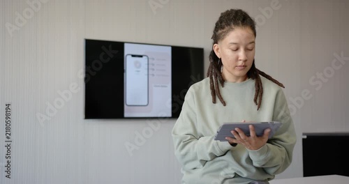 African American nonbinary sitting stool tapping tablet being triggered by wall UI pausing thinking