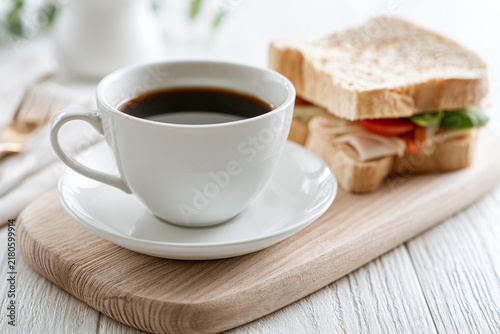 White cup of black coffee on a saucer next to a sandwich on a wooden cutting board