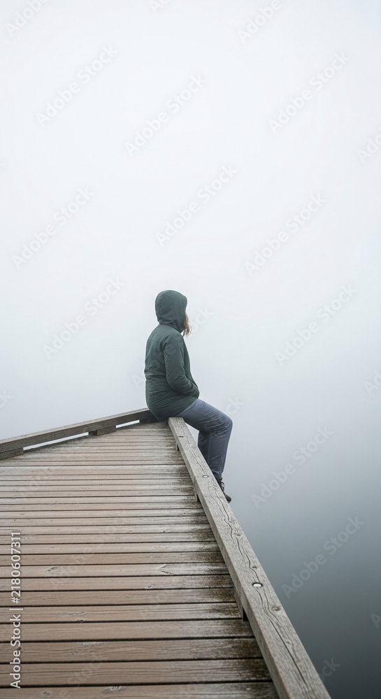Fototapeta premium A person in a hooded jacket sits on a wooden pier, looking into the dense fog symbolizing contemplation and isolation