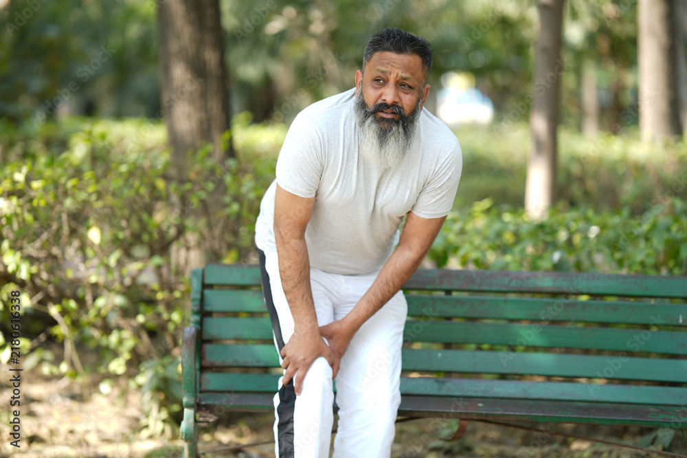 Fototapeta premium Indian senior man suffering from knee pain at park. Pain In The Elderly, Health care. Bearded man painful on knee.