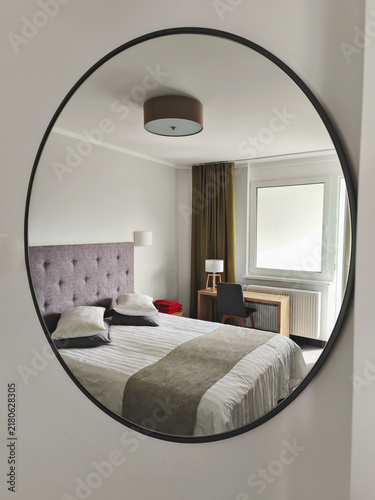 Oval wall mirror reflecting modern bedroom with double bed, upholstered headboard, window, desk and soft neutral decor