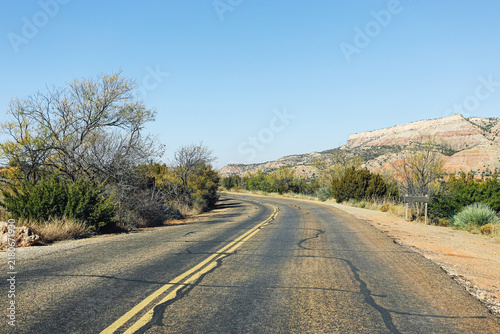 Fotografie Empty asphalt road curves through dry scrubland toward rocky hills under a clear blue sky