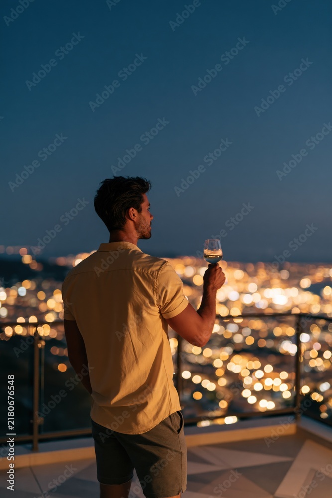 Obraz premium Man on balcony holding a wine glass at night with city skyline background for rooftop lifestyle banner and background design