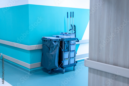 A cleaning trolley with mops, buckets and a laundry bag stands in the corner of a hospital corridor. Turquoise walls rise above the wipe clean floor. Sanitary service in a new modern hospital