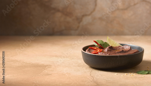 A bowl contains creamy Mexican black bean dip with slices of lime and red chili on top. The setting has a simple and minimalist design. It features a smooth surface