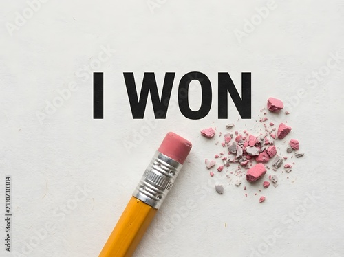Pencil erasing the text i won on a white paper surface leaving pink eraser crumbs behind
