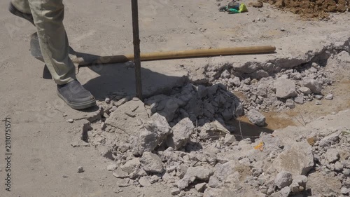 A cropped view shows a construction worker using a long metal pry bar to loosen broken concrete slabs during road repair, with exposed wire, wet ground and scattered tools creating workplace hazards.