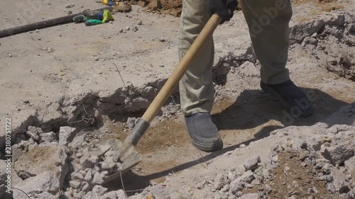 A cropped construction worker shovels heavy concrete rubble from demolished pavement while exposed wire and unstable debris create workplace hazards during outdoor road repair.
