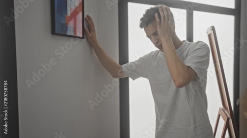 Young man with hand on forehead, leaning on wall and supporting himself by an easel in studio; distress dizziness sickness.