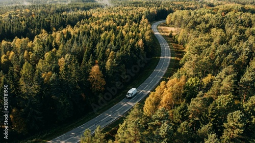 Aerial view of a white van traveling on a winding road through a vibrant autumn forest
