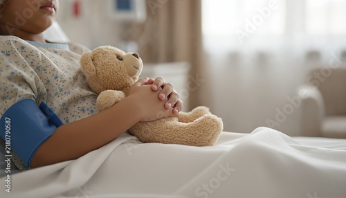 Young girl resting in a bright pediatric hospital bed with a blood pressure cuff while holding a soft plush toy for comfort