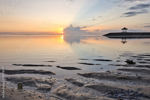 Obraz na płótnie Distinctive sand patterns at low tide on Sanur beach during a serene golden sunrise, with Mount Agung volcano silhouette and traditional gazebos in Bali