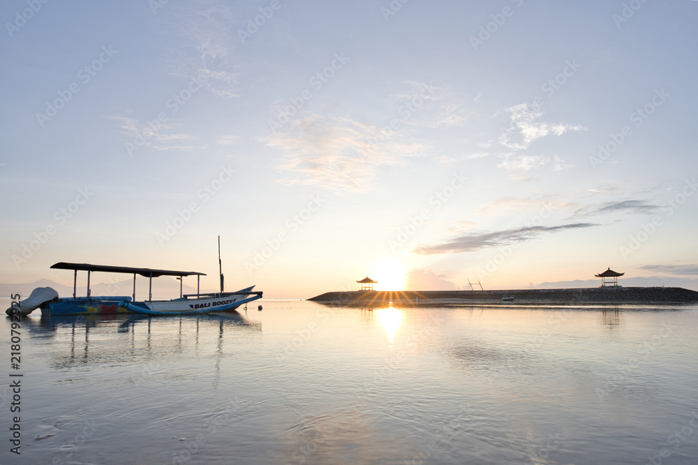 Fototapeta premium Distinctive sand patterns at low tide on Sanur beach during a serene golden sunrise, with Mount Agung volcano silhouette and traditional gazebos in Bali.