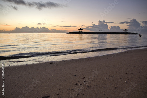 Peaceful morning landscape of Sanur beach featuring traditional gazebos on a stone breakwater jetty under a beautiful dramatic cloud sky at sunrise in Bali, Indonesia.