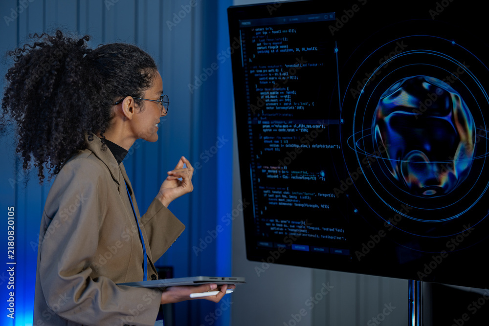 Obraz premium Black female cybersecurity analyst with curly hair and glasses types at a keyboard while code and data fill large screens behind her.