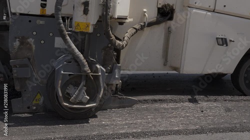 A cold milling machine removes old asphalt, showing its hydraulic wheel drive, hoses and grooved pavement in a close side view during road repair.