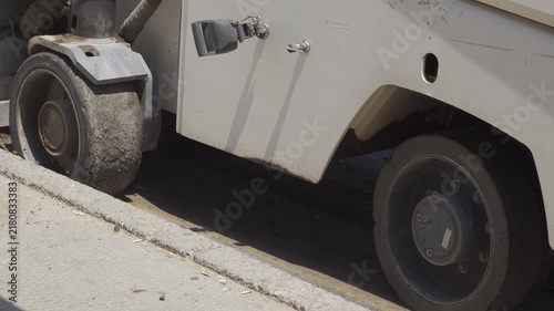 Side close-up of an asphalt milling machine moving beside a curb, showing its large wheel, white chassis and freshly milled pavement during road resurfacing.