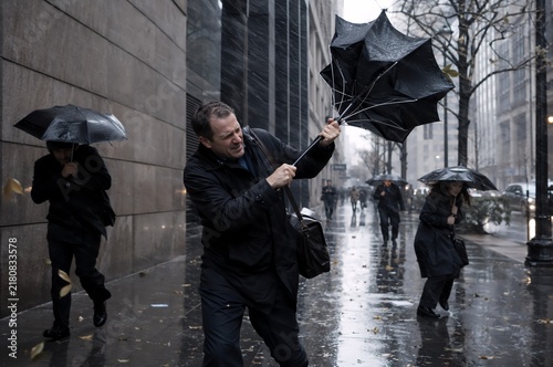 Man struggling with broken umbrella in strong wind and rain on city street. Businessman with bag commuting to work in bad weather. People walking under umbrellas during autumn storm in downtown.