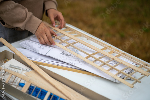 Teenager assembling wooden model airplane wing from balsa parts on table with blueprints. Hobby of aircraft modeling and engineering education for kids. Handmade construction and creativity concept.