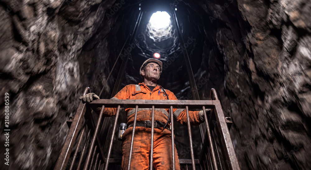 Fototapeta premium Low angle view of a miner in protective gear looking upwards through the metal bars of a hoist elevator inside a rocky underground mine shaft