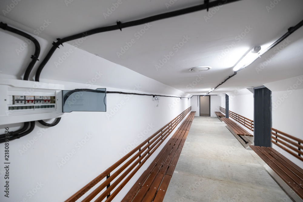 Fototapeta premium Underground bomb shelter corridor with white walls, long wooden benches, electrical panel, conduit pipes running along the ceiling, fluorescent lights, concrete floor, leading to a closed metal door