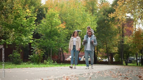 Family of three walking outdoors in park during autumn. man carries child on his shoulders while accompanied by woman all moving along path with fallen leaves and green trees surrounding