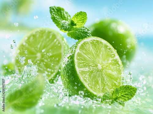 A sliced fresh green lime rests splashing in clear blue water, paired with vibrant mint leaves, whole limes blurred in soft‑focus tropical background.