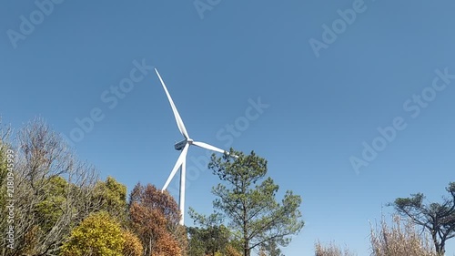 A majestic white wind turbine rises behind colorful treetops under a clear blue sky, symbolizing hope and sustainable green energy. Perfect for ecology concepts. No people.
