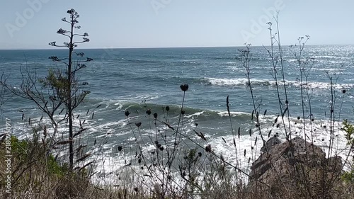 A serene coastal view of shimmering blue ocean waves crashing below a wild cliffside. Nobody is present, evoking a deep sense of peaceful solitude and raw, untouched natural beauty.