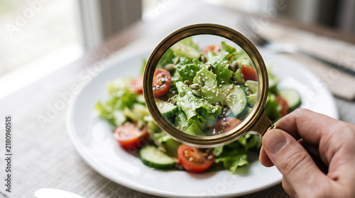 Woman hand examining fresh vegetable salad through magnifying glass. Concept of orthorexia and obsessive control of food intake. Disordered eating and unhealthy relationship with nutrition.