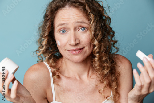 Obraz na plátně A middle aged woman with natural skin and curly hair looks surprised while holding cosmetic boxes in a studio with a blue background, conveying product browsing and authentic unretouched beauty