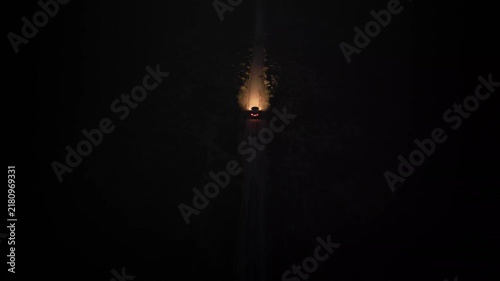 A single vehicle drives down an unpaved road in the desert at night with headlights illuminating the dirt path ahead, shown from an elevated, following view in real time.