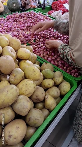 A person in floral sleeves selects small red shallots from a green crate at a market, next to a large pile of yellow potatoes, with garlic and pumpkins visible in the background.