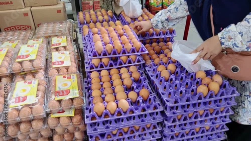 A person arranges brown chicken eggs in plastic trays at a crowded supermarket aisle displayed alongside packaged cartons and stacked cardboard boxes.
