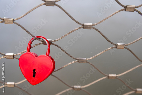 Red heart-shaped padlock attached to a wire mesh metal fence railing over water, love lock tradition.