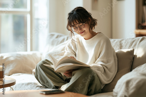 Obraz na płótnie スマホを机に置いて眼鏡をかけリネンのソファで本を読む日本人女性、デジタルデトックス