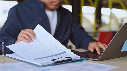 Businessman performs financial analysis reviewing paper document, conducting market analysis beside modern laptop as data analysis clarifies key document details.