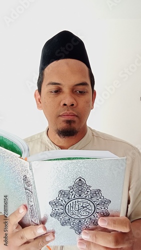 Bild auf Leinwand A Muslim man wearing a traditional black cap and beige attire is reading the Qur