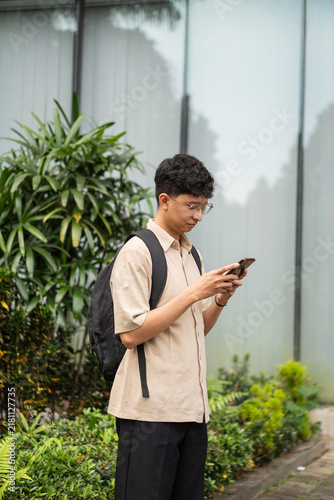 Принти на полотні A young man stands outdoors, engrossed in his smartphone while surrounded by lush greenery