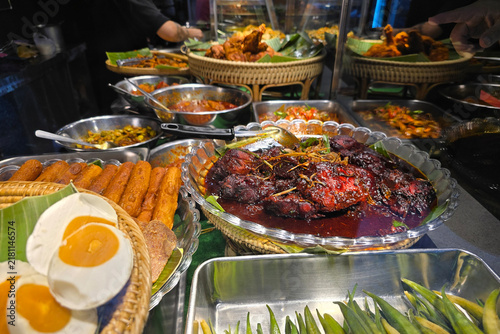Obraz na plátně Close-up display of traditional Malaysian nasi lemak side dishes, featuring spicy chicken in red gravy (ayam masak merah), salted eggs, lady's fingers, and sambal varieties at a food stall
