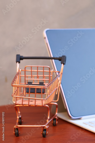Miniature shopping cart in front of an open tablet with blank mockup screen on wooden table, online shopping and digital commerce concept.