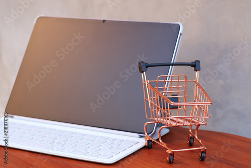 Miniature shopping cart in front of an open tablet with blank mockup screen on wooden table, online shopping and digital commerce concept.